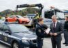 Odvzem vozila, zakaj naknadna „prodaja” avta ne deluje in kaj lahko naredite drugače Police car in the foreground as a tow truck hauls an orange sports car; a policewoman hands paperwork to a suited man while another officer talks on a mobile phone nearby.