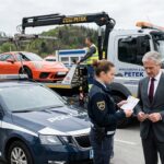 Odvzem vozila, zakaj naknadna „prodaja” avta ne deluje in kaj lahko naredite drugače Police car in the foreground as a tow truck hauls an orange sports car; a policewoman hands paperwork to a suited man while another officer talks on a mobile phone nearby.