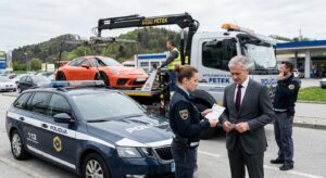 Police car in the foreground as a tow truck hauls an orange sports car; a policewoman hands paperwork to a suited man while another officer talks on a mobile phone nearby.