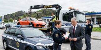 Odvzem vozila, zakaj naknadna „prodaja” avta ne deluje in kaj lahko naredite drugače Police car in the foreground as a tow truck hauls an orange sports car; a policewoman hands paperwork to a suited man while another officer talks on a mobile phone nearby.