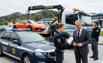 Odvzem vozila, zakaj naknadna „prodaja” avta ne deluje in kaj lahko naredite drugače Police car in the foreground as a tow truck hauls an orange sports car; a policewoman hands paperwork to a suited man while another officer talks on a mobile phone nearby.