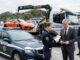 Odvzem vozila, zakaj naknadna „prodaja” avta ne deluje in kaj lahko naredite drugače Police car in the foreground as a tow truck hauls an orange sports car; a policewoman hands paperwork to a suited man while another officer talks on a mobile phone nearby.